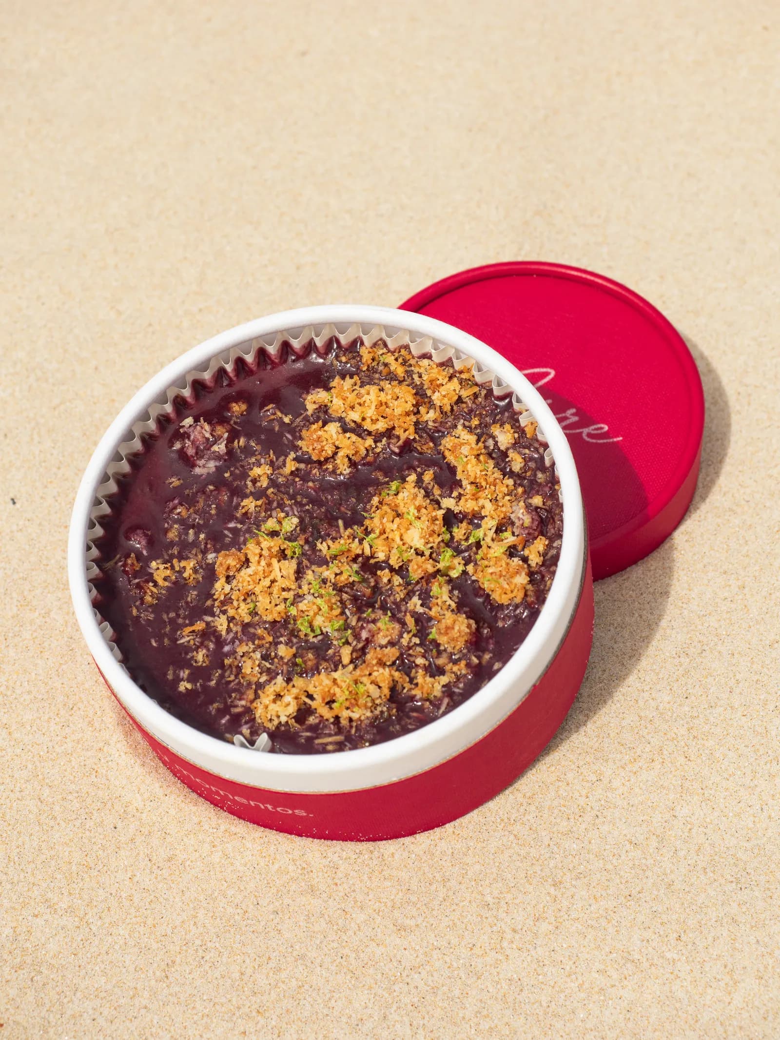 Cheesecake de açaí, morada y con crumble dorado por encima, en su tarrina roja de Aire sobre la arena.