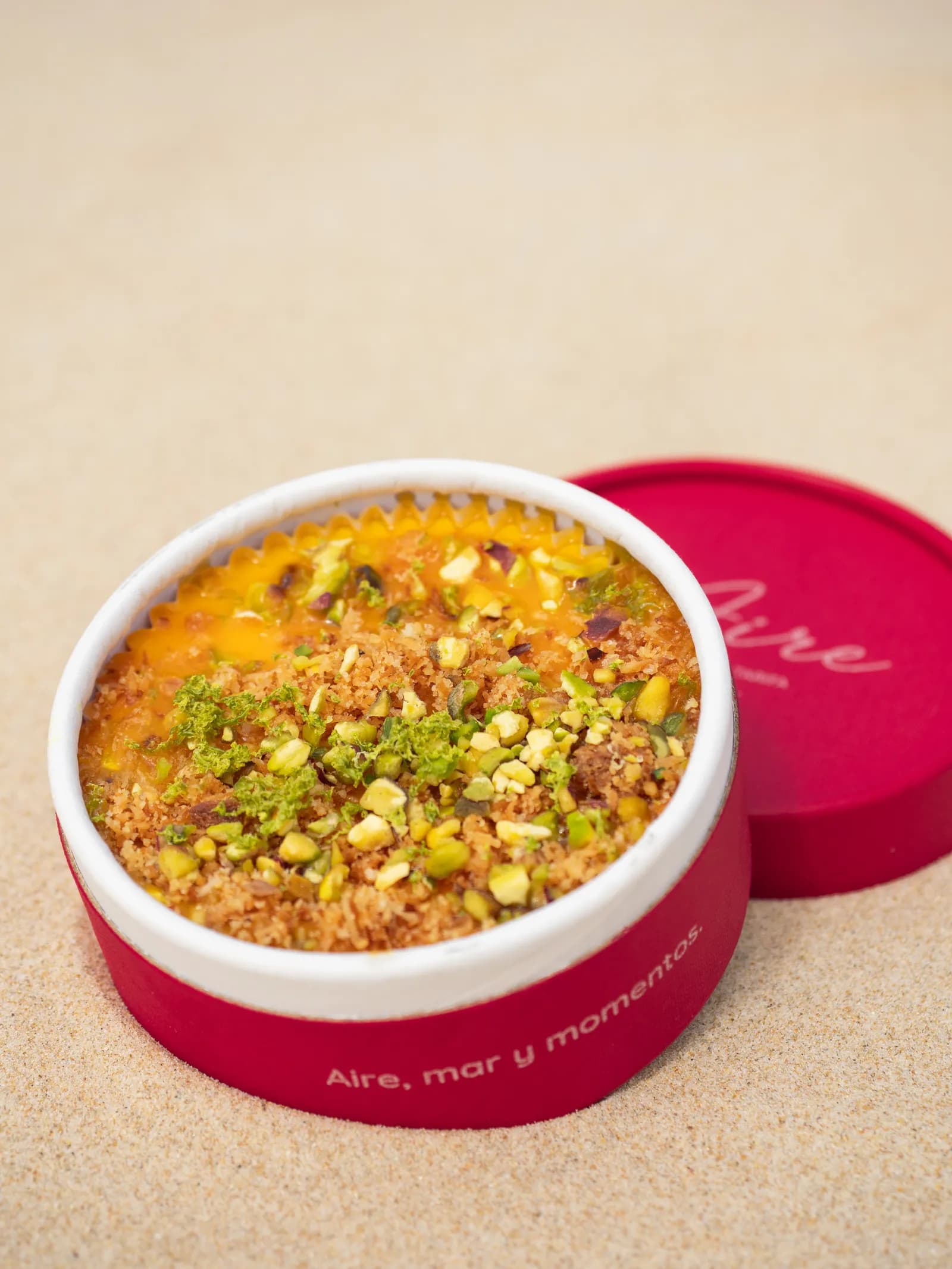 Cheesecake de mango y maracuyá con crumble y pistacho picado por encima, en su tarrina roja de Aire sobre la arena.